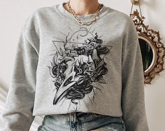 Op de afbeelding: Een grijs gecropped sweatshirt met een zwart-witte grafische print van een schedel met een vogel erop. De grafiek is omgeven door een geometrisch ontwerp. Het sweatshirt wordt gedragen door een persoon met een witte achtergrond.