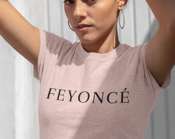 Può includere: Una donna indossa una maglietta rosa con la scritta "FEYONCÉ" stampata sul davanti. Indossa pantaloni neri e ha i capelli ricci.