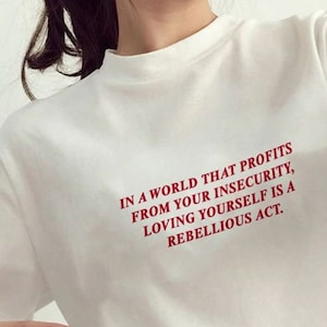 Może przedstawiać: Biała koszulka z czerwonym napisem: "IN A WORLD THAT PROFITS FROM YOUR INSECURITY, LOVING YOURSELF IS A REBELLIOUS ACT."