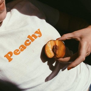 Op de afbeelding: Een wit T-shirt met het woord "Peachy" bedrukt in oranje letters. Een persoon houdt een gehalveerde perzik in zijn hand.