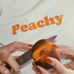 Op de afbeelding: Een persoon snijdt een perzik doormidden met een zilveren mes. De perzik is rijp en sappig, met een helder oranje vruchtvlees. De persoon draagt een wit shirt met het woord "Peachy" erop gedrukt in oranje letters.