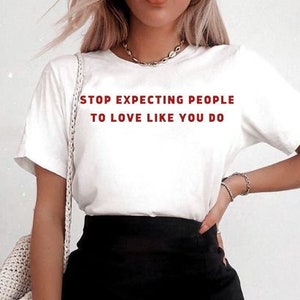 Puede incluir: Una camiseta blanca con el texto "STOP EXPECTING PEOPLE TO LOVE LIKE YOU DO" en letras rojas. La modelo lleva una minifalda negra con una abertura lateral.