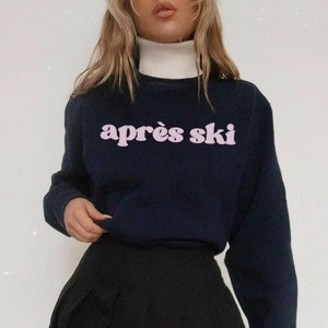 Op de afbeelding: Een donkerblauwe trui met een roze "apr&egrave;s ski"-print. De trui wordt gecombineerd met een zwarte geplooide rok.