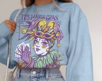 May include: A light blue sweatshirt featuring a graphic of a jester holding playing cards. The graphic is in shades of purple, yellow, green, and blue. The text "IT'S MARIDIGRAS" is printed on the sweatshirt.