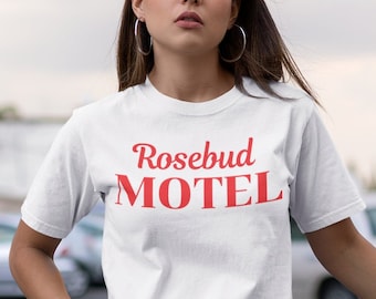 May include: A woman wearing a white t-shirt with the text "Rosebud Motel" in red lettering. She is wearing blue jeans with distressed details and is standing in a car park.