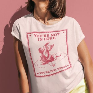 Pu&ograve; includere: Una maglietta bianca con una grafica rosa di un cupido che beve da una bottiglia con il testo "You're not in love, you're just drunk".