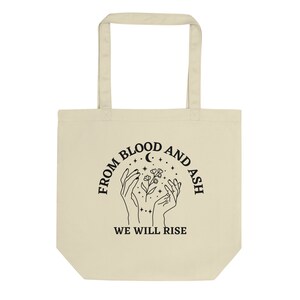 May include: A beige tote bag with black text and a graphic design. The text reads "From Blood and Ash We Will Rise". The graphic design features two hands holding a flower with a crescent moon and stars in the background.