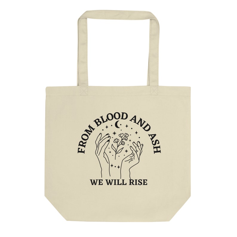 May include: A beige tote bag with black text and a graphic design. The text reads "From Blood and Ash We Will Rise". The graphic design features two hands holding a flower with a crescent moon and stars in the background.