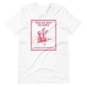 Pu&ograve; includere: Maglietta bianca con una grafica rossa di un cupido che beve da una bottiglia con il testo "You're not in love. You're just drunk."