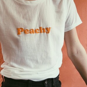 Op de afbeelding: Een wit T-shirt met het woord "Peachy" in oranje letters.