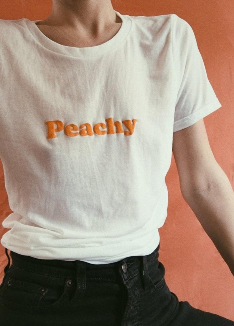 Op de afbeelding: Een wit T-shirt met het woord "Peachy" in oranje letters.