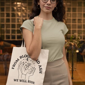 May include: A white tote bag with black text that reads "From Blood and Ash We Will Rise". The text is in a circle with a crescent moon and stars. Two hands are holding a flower with leaves.