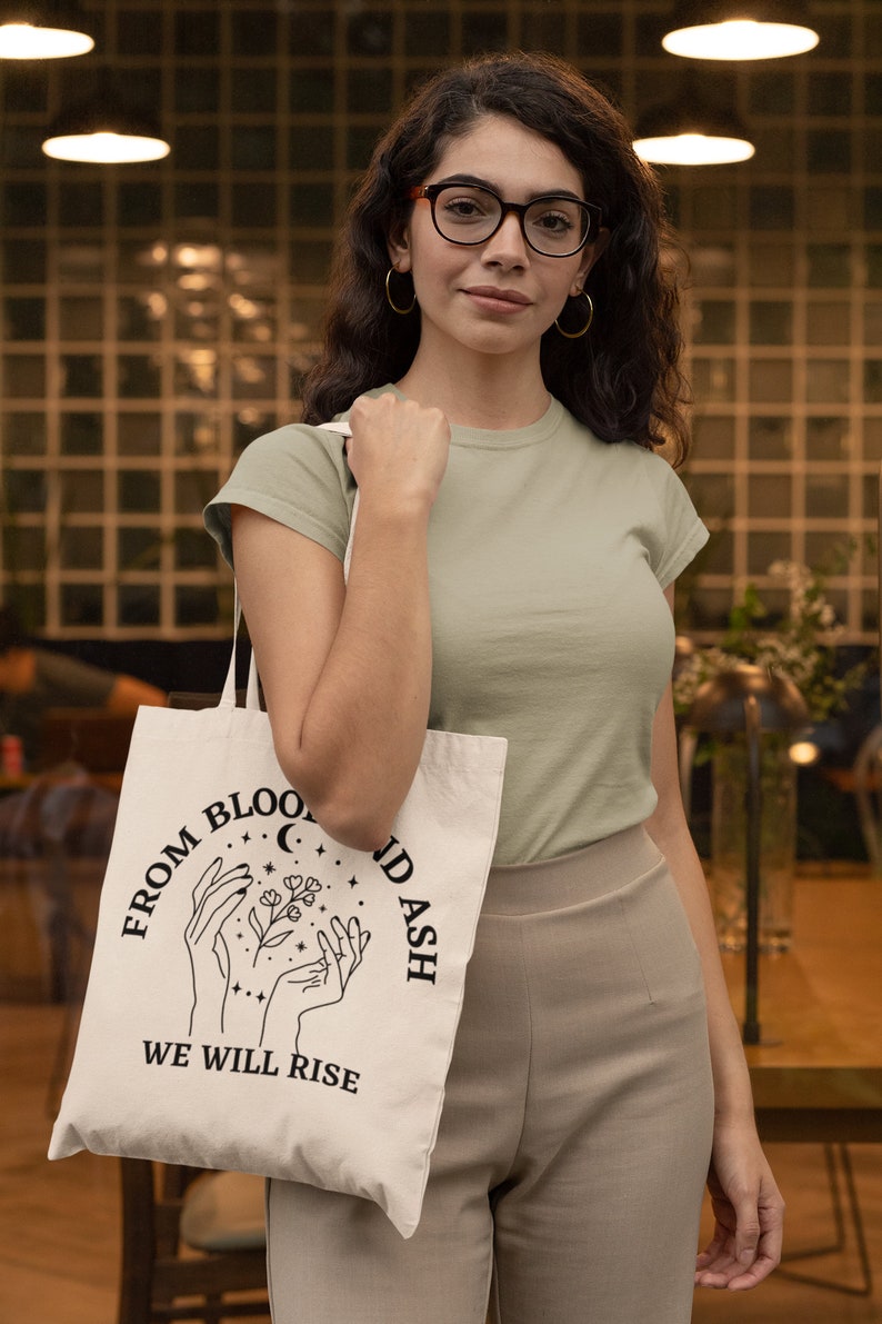 May include: A white tote bag with black text that reads "From Blood and Ash We Will Rise". The text is in a circle with a crescent moon and stars. Two hands are holding a flower with leaves.