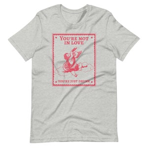Pu&ograve; includere: Una t-shirt grigia melange con una grafica rosa di un cupido che beve da una bottiglia. La grafica &egrave; in stile vintage ed &egrave; circondata da un bordo rosa con cuori. Il testo sulla grafica dice "You're not in love. You're just drunk."
