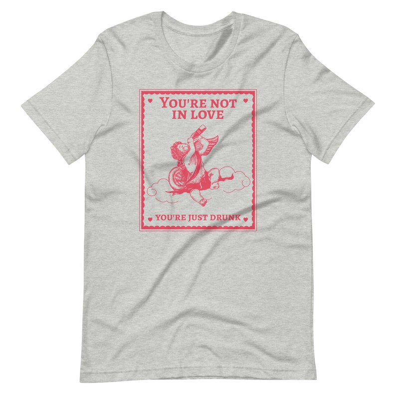 Pu&ograve; includere: Una t-shirt grigia melange con una grafica rosa di un cupido che beve da una bottiglia. La grafica &egrave; in stile vintage ed &egrave; circondata da un bordo rosa con cuori. Il testo sulla grafica dice "You're not in love. You're just drunk."