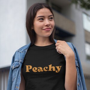 Op de afbeelding: Een zwart T-shirt met het woord "Peachy" gedrukt in een retro oranje lettertype.
