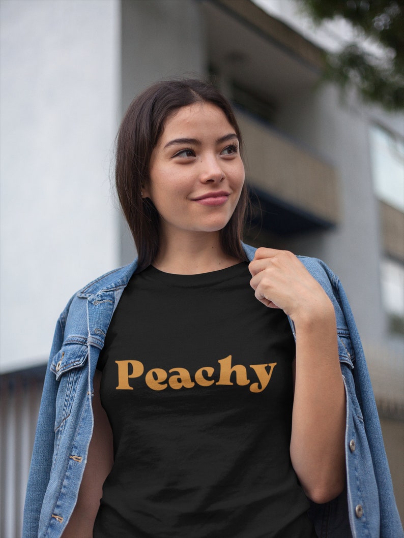 Op de afbeelding: Een zwart T-shirt met het woord "Peachy" gedrukt in een retro oranje lettertype.