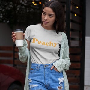 Op de afbeelding: Een vrouw draagt een wit T-shirt met het woord "Peachy" in oranje gedrukt. Ze draagt ook een lichtgroene cardigan en een blauwe denim short.