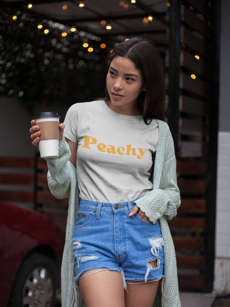 Op de afbeelding: Een vrouw draagt een wit T-shirt met het woord "Peachy" in oranje gedrukt. Ze draagt ook een lichtgroene cardigan en een blauwe denim short.