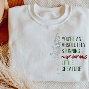 Op de afbeelding: Witte trui met een zwarte lijntekening van een mes met bloemen erop. De tekst "You're an absolutely stunning murderous little creature" staat op de trui gedrukt.