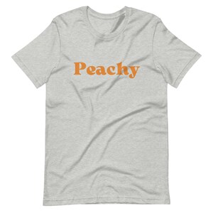 Op de afbeelding: Een grijs t-shirt met het woord "Peachy" in oranje gedrukt.