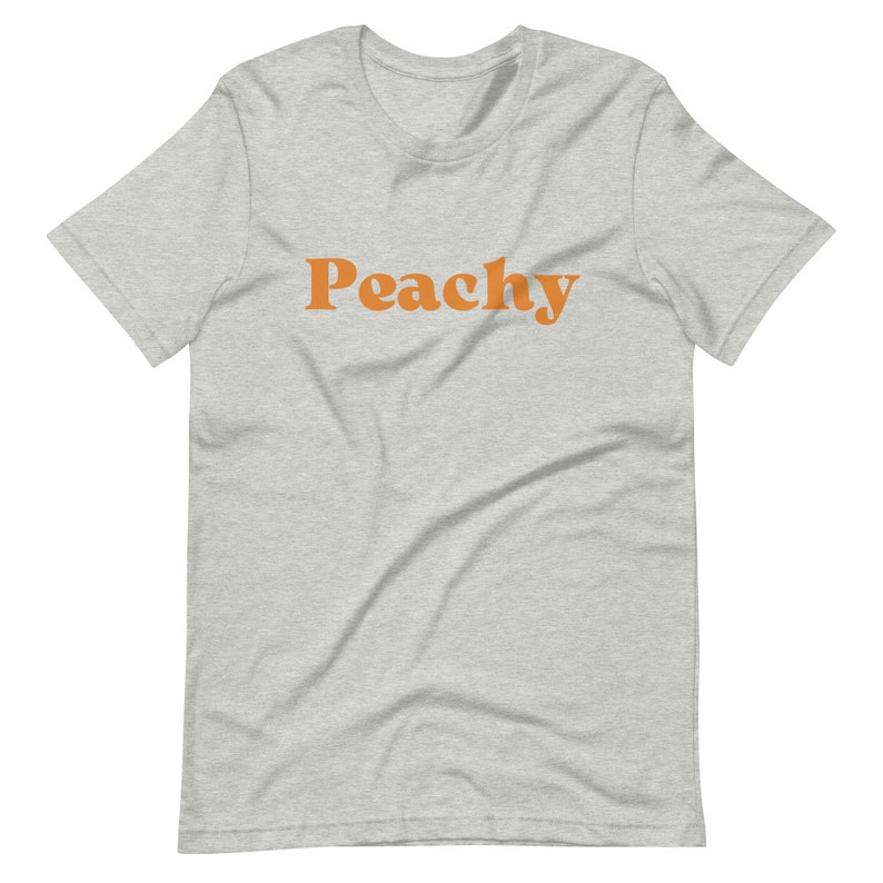 Op de afbeelding: Een grijs t-shirt met het woord "Peachy" in oranje gedrukt.