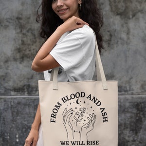 May include: A beige tote bag with black text and a graphic of two hands holding a flower with a crescent moon and stars. The text reads "From Blood and Ash We Will Rise."