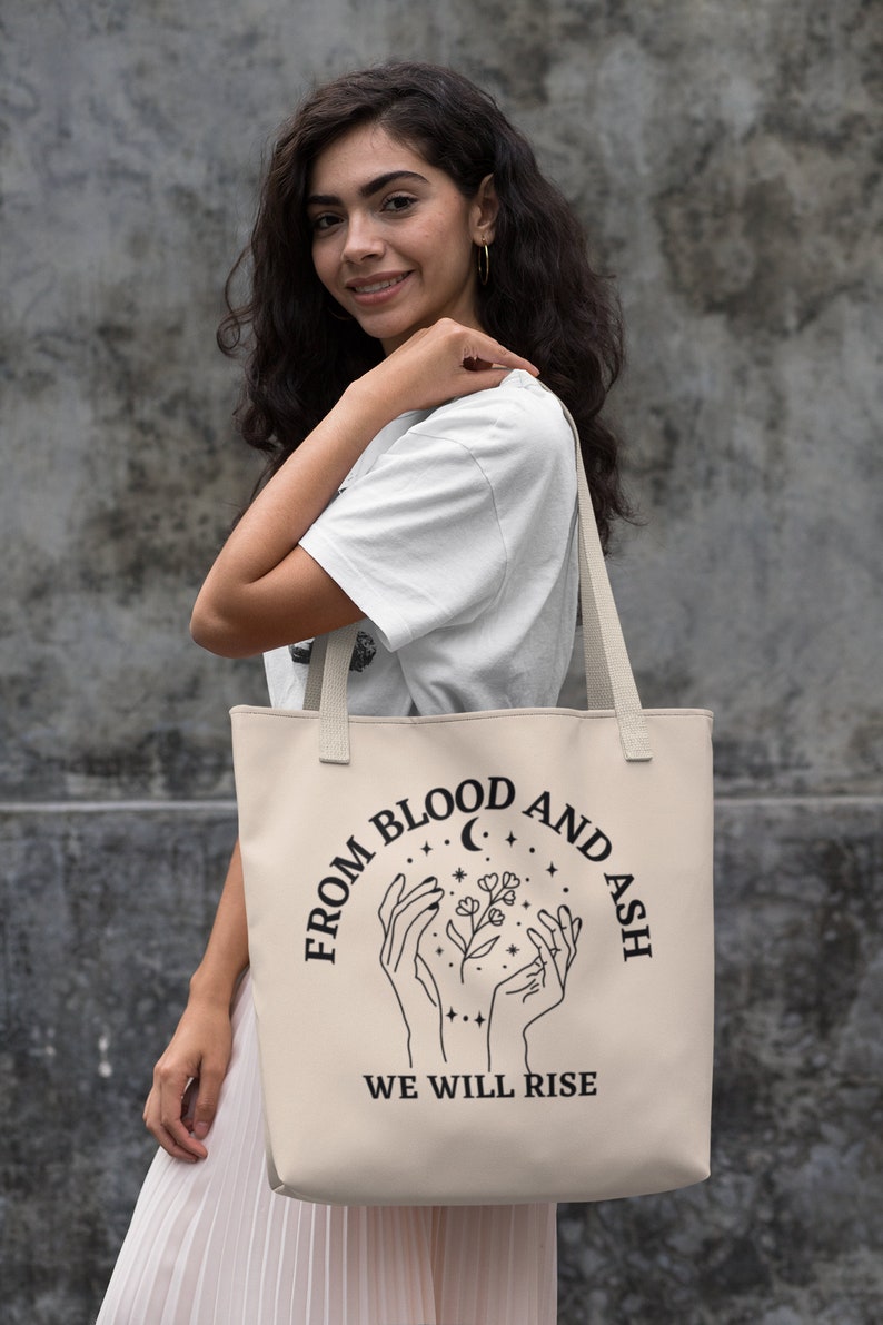 May include: A beige tote bag with black text and a graphic of two hands holding a flower with a crescent moon and stars. The text reads "From Blood and Ash We Will Rise."
