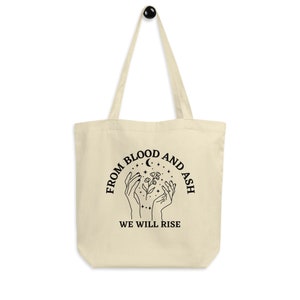 May include: A beige tote bag with black text and a graphic design. The text reads "From Blood and Ash We Will Rise". The graphic design features two hands holding a flower with stars and a crescent moon.