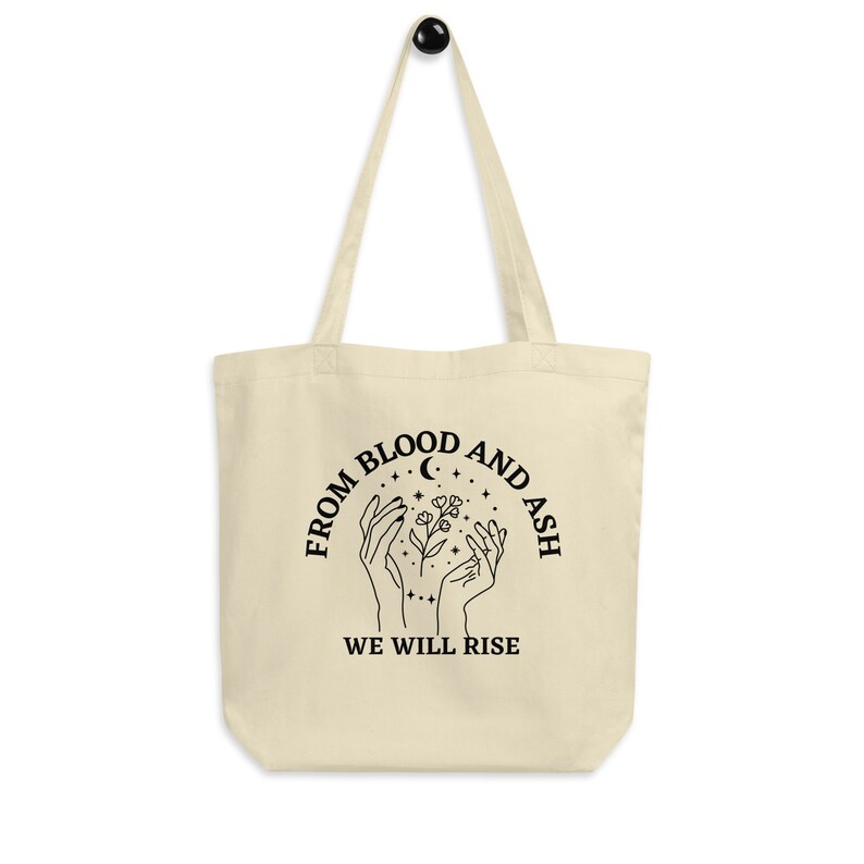 May include: A beige tote bag with black text and a graphic design. The text reads "From Blood and Ash We Will Rise". The graphic design features two hands holding a flower with stars and a crescent moon.
