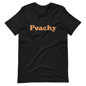Op de afbeelding: Zwart T-shirt met het woord "Peachy" in oranje gedrukt.