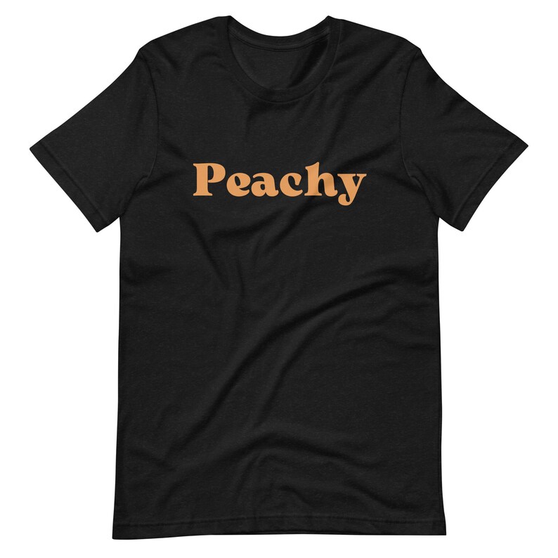 Op de afbeelding: Zwart T-shirt met het woord "Peachy" in oranje gedrukt.