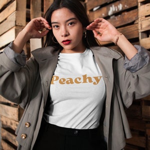 Op de afbeelding: Een vrouw draagt een wit T-shirt met het woord "Peachy" erop gedrukt in een retro oranje lettertype. Ze draagt een grijze trenchcoat en een zwarte broek.