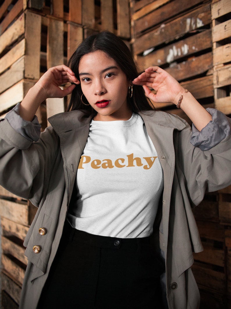 Op de afbeelding: Een vrouw draagt een wit T-shirt met het woord "Peachy" erop gedrukt in een retro oranje lettertype. Ze draagt een grijze trenchcoat en een zwarte broek.