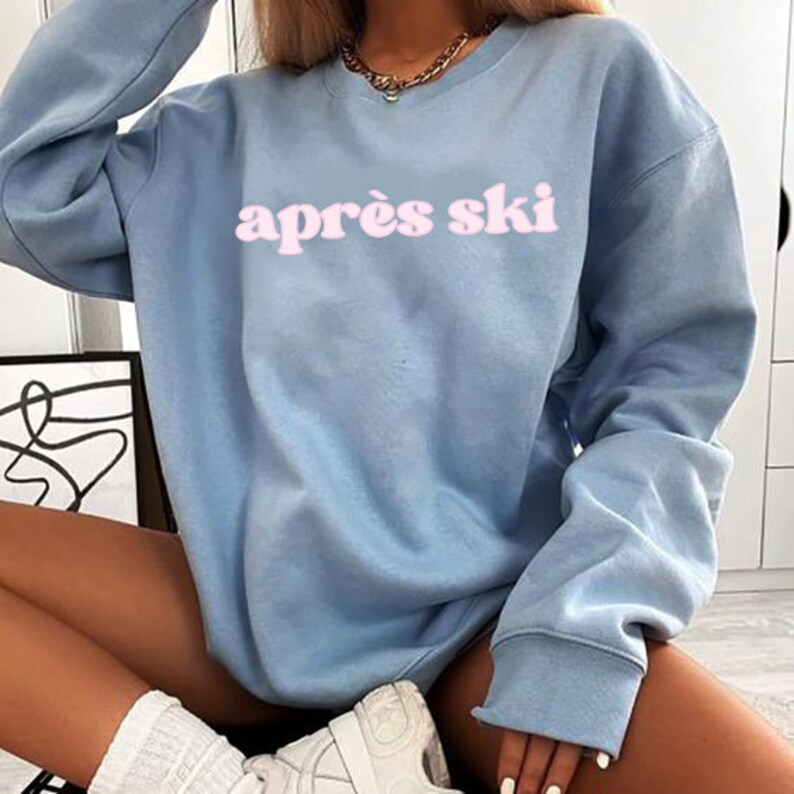 Op de afbeelding: Een lichtblauw sweatshirt met de tekst "apr&egrave;s ski" in roze letters.
