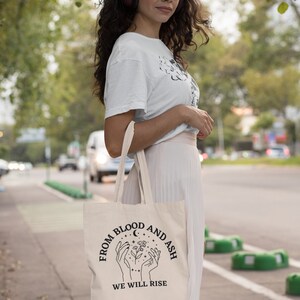 May include: A white tote bag with black text and a graphic design. The text reads "From Blood and Ash We Will Rise".