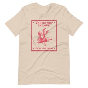 Pu&ograve; includere: Una t-shirt beige con una grafica rosa di un cupido che beve da una bottiglia. La grafica &egrave; in stile vintage e ha il testo "You're not in love. You're just drunk."
