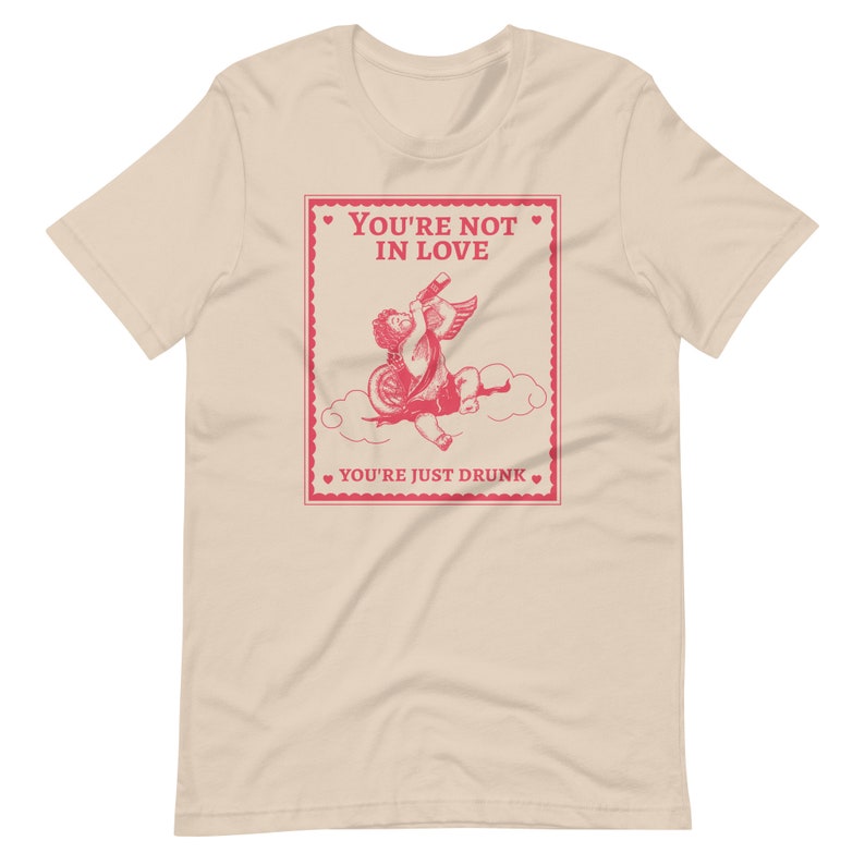 Pu&ograve; includere: Una t-shirt beige con una grafica rosa di un cupido che beve da una bottiglia. La grafica &egrave; in stile vintage e ha il testo "You're not in love. You're just drunk."