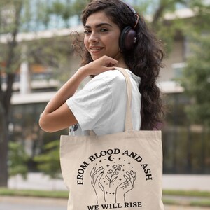 May include: A beige tote bag with black text that reads "From Blood and Ash We Will Rise". The text is in a circle with a crescent moon and stars. There are two hands holding a flower in the center of the circle.