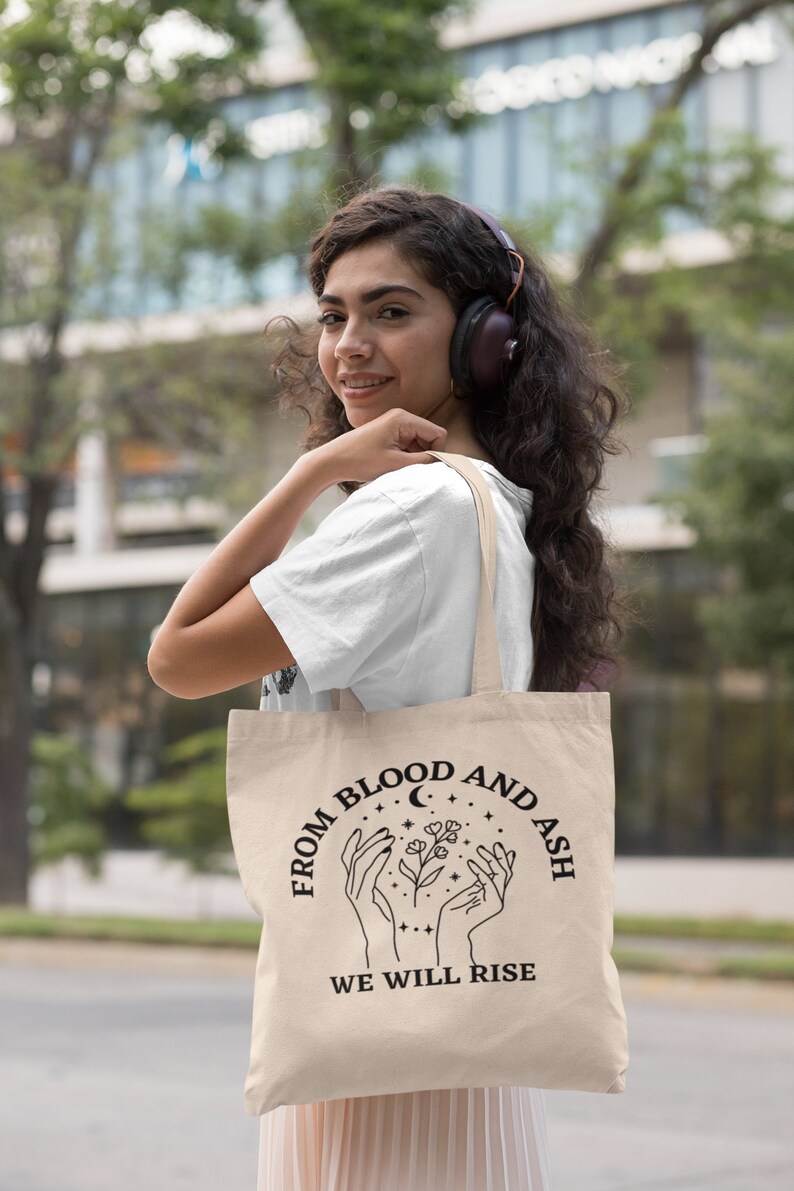 May include: A beige tote bag with black text that reads "From Blood and Ash We Will Rise". The text is in a circle with a crescent moon and stars. There are two hands holding a flower in the center of the circle.