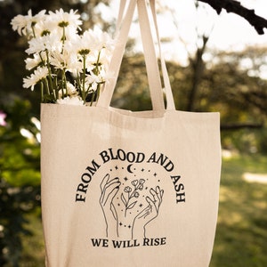 May include: A beige tote bag with black text and a graphic of two hands holding a flower with stars and a crescent moon. The text reads "From Blood and Ash We Will Rise."