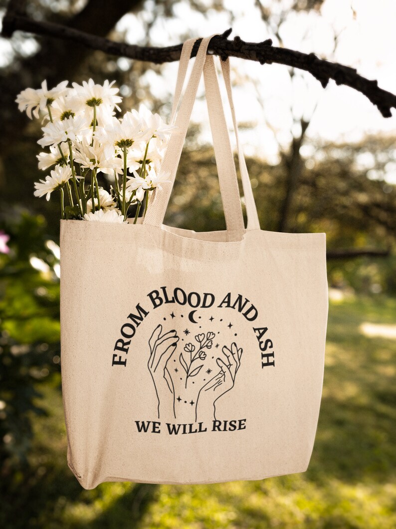 May include: A beige tote bag with black text and a graphic of two hands holding a flower with stars and a crescent moon. The text reads "From Blood and Ash We Will Rise."