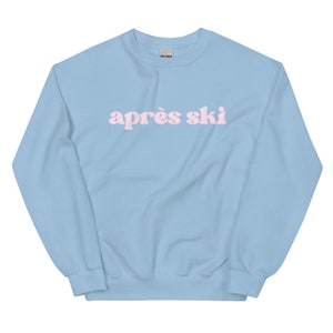 Op de afbeelding: Een lichtblauw sweatshirt met de tekst "apr&egrave;s ski" in roze letters.