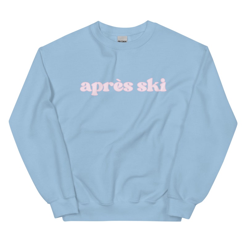 Op de afbeelding: Een lichtblauw sweatshirt met de tekst "apr&egrave;s ski" in roze letters.