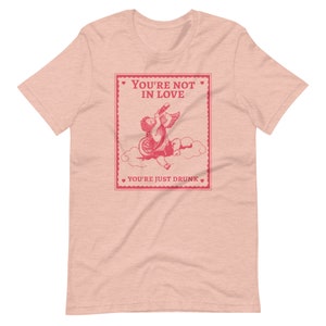 Pu&ograve; includere: Maglietta rosa con una grafica rossa di un cupido che beve da una bottiglia con il testo "You're not in love. You're just drunk."