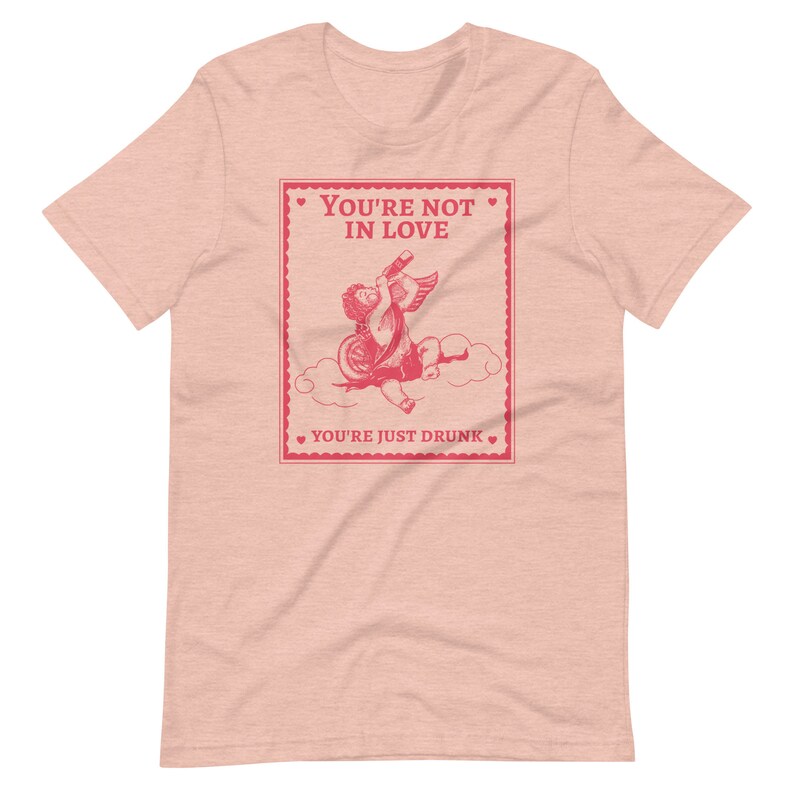 Pu&ograve; includere: Maglietta rosa con una grafica rossa di un cupido che beve da una bottiglia con il testo "You're not in love. You're just drunk."