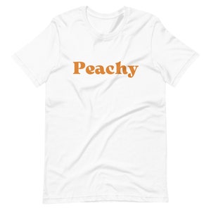 Op de afbeelding: Wit T-shirt met het woord "Peachy" in oranje op de voorkant.
