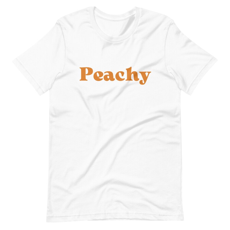 Op de afbeelding: Wit T-shirt met het woord "Peachy" in oranje op de voorkant.