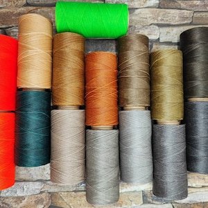 Puede incluir: Surtido de carretes de hilo encerado en varios colores, incluyendo naranja, verde, marrón, beige y gris. Los carretes están dispuestos en una fila sobre un fondo de piedra.