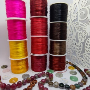 Può includere: Cinque bobine di cordoncino elastico in diversi colori: rosa, rosso, nero, marrone e rosso scuro. Le bobine sono disposte in fila su una superficie bianca. Ci sono anche alcune perline e pietre sparse intorno alle bobine.